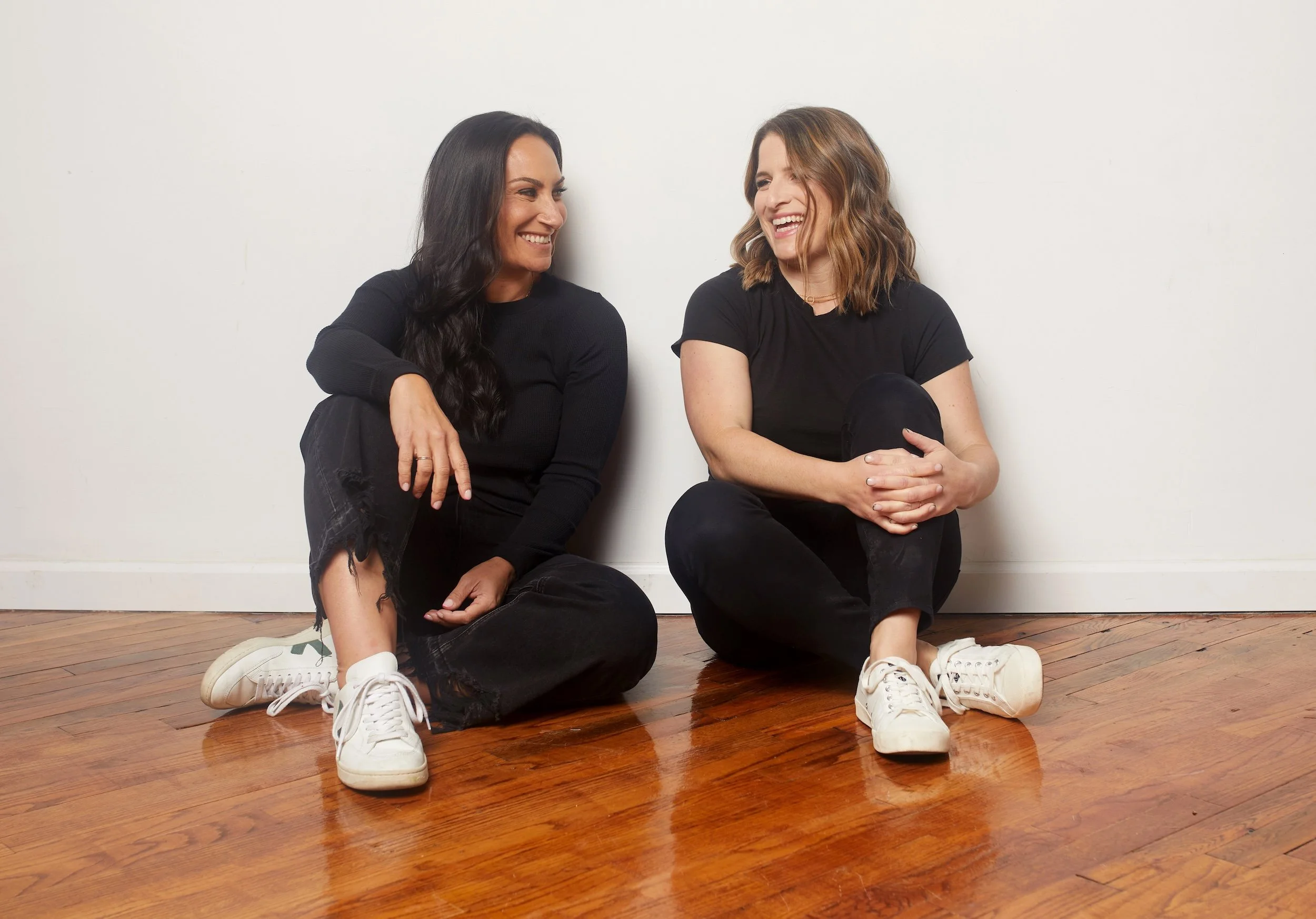 Jenna Shapiro and Stacy Berger of Work Better Together, laughing, seated on a wooden floor against a white wall